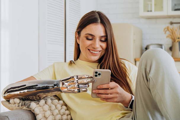 A happy woman with a bionic prosthetic arm sits on the couch with a phone and communicates online with friends or surfs the Internet or makes online purchases - Photo, Image