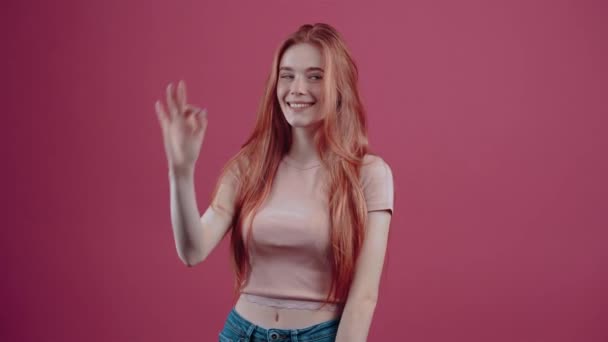 The young redhead with a beautiful smile, shows with her hand the gesture well, smiling happily at the room. 20-year-old freckled hipster teenager in a pink casual T-shirt, isolated on a pink - Footage, Video