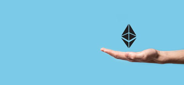 Αρσενικό χέρι που κρατά ένα εικονίδιο Ethereum σε μπλε φόντο. Ethereum και cryptocurrency επένδυση έννοια. ανταλλαγή, εμπορία, μεταφορά και επένδυση τεχνολογίας blockchain - Φωτογραφία, εικόνα