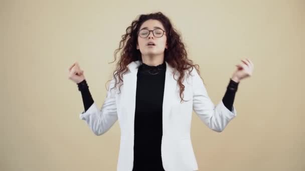 The young woman with calm and calm curly hair holds her hands sideways in the form of yoga, relaxing. Young hipster in gray jacket and white shirt, with glasses posing isolated on beige background in - Footage, Video