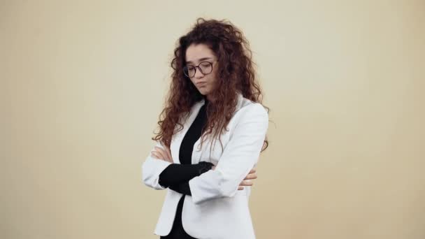 Upset young woman with curly hair, holds her hands in the middle and looks contemptuously into the room. Young hipster in gray jacket and white shirt, with glasses posing isolated on beige background - Footage, Video