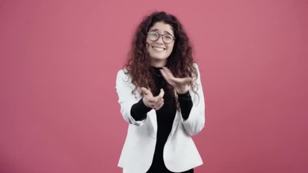 The young adult with curly hair gestures with her hands in front of her, smiling ironically. Young hipster in white jacket and black shirt, with glasses posing isolated on pink background in the - Footage, Video