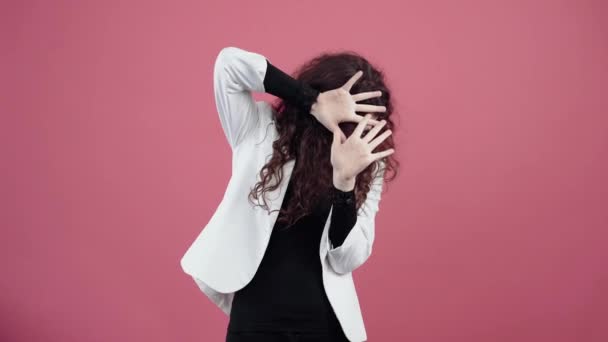 Frightened young woman with curly hair, she keeps her hands in cotton wool hiding behind her palms. Young hipster in white jacket and black shirt, with glasses posing isolated on pink background in - Footage, Video