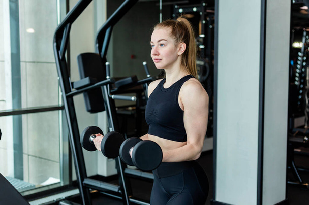 A young girl in black sportswear is engaged in the gym with dumbbells. Fitness club concept - Фото, изображение