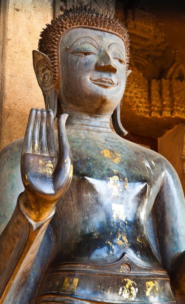 The bronze Buddha image in standing posture wiht lifted right hand in front of the sandstone handcrafted church. - Photo, Image