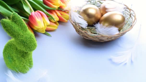 Canasta de huevos de Pascua. Huevo dorado en cesta con tulipanes primaverales, plumas blancas sobre fondo azul pastel en feliz decoración de Pascua. Decoración tradicional en luz solar - Metraje, vídeo
