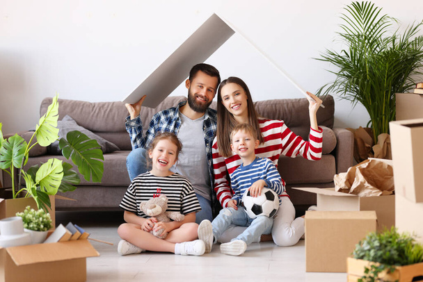 Cheerful parents and kids holding house roof over head and smiling for camera while sitting near couch and belongings in new flat - Фото, изображение