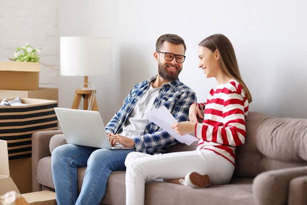 Onnellinen nuori pari kannettavan tietokoneen ja paperit istuu sohvalla huoneessa pakattu laatikot ja keskustella vakuutussopimuksen muuton jälkeen uuteen asuntoon - Valokuva, kuva
