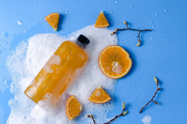 Shampooing, gel douche, savon liquide orange avec des ingrédients naturels dans un flacon transparent sur fond bleu en mousse savonneuse avec des morceaux d'orange, espace de copie plat - Photo, image