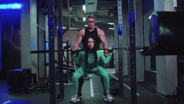 Woman in sports costume trains her butt with dumbbell on her shoulders under the supervision of a trainer - Footage, Video