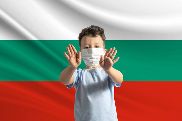 Μικρό λευκό αγόρι με προστατευτική μάσκα στο φόντο της σημαίας της Βουλγαρίας. Κάνει στοπ με τα χέρια, μένει σπίτι Βουλγαρία. - Φωτογραφία, εικόνα