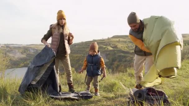 Slowmo medium shot of young woman, man and cute 5-year-old boy setting up camp on top of hill overlooking lake on sunny day - Footage, Video