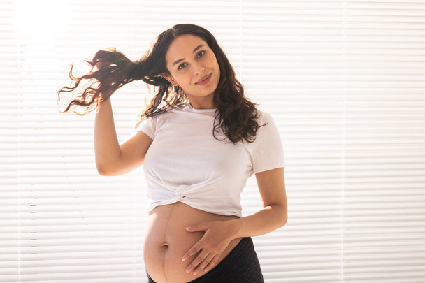 Улыбающаяся молодая красивая беременная женщина, трогающая живот, волосы и радующаяся. Концепция здоровья и размышления о будущем в ожидании ребенка. Copyspace - Фото, изображение