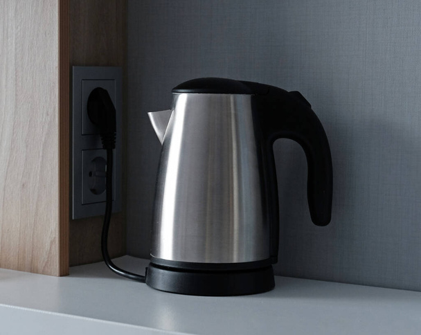 Silberner Wasserkocher aus Metall zum Kochen von Wasser und zum Kochen von Tee oder Kaffee auf einem Tisch. Haushaltsküchengeräte für Heißgetränke. Wasserkocher aus rostfreiem Stahl wird an eine Steckdose angeschlossen. - Foto, Bild