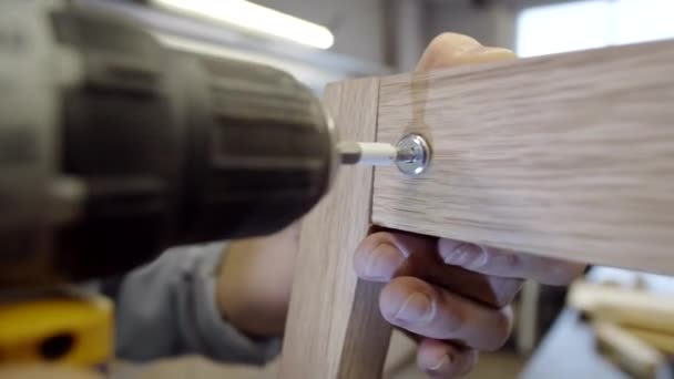 Κοντινό πλάνο των χεριών του ανώνυμου αρσενικού ξυλουργού οδήγηση σε βίδα με ηλεκτρικό τρυπάνι, ενώ κάνοντας στερεά έπιπλα από ξύλο στο εργαστήριο - Πλάνα, βίντεο