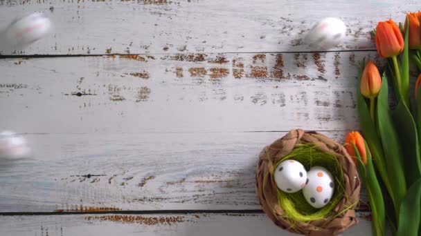 Vídeo 4k. Huevos multicolores de Pascua rodando sobre mesa de madera blanca con tulipanes. Composición tradicional de primavera Elementos de Pascua flores y huevos para primavera. Concepto de decoraciones de primavera - Metraje, vídeo
