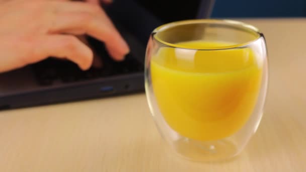 Szkło z sokiem pomarańczowym na pierwszym planie. Kobieta pracuje za laptopem w tle i okresowo podnosi filiżankę drinka jego ręką.. - Materiał filmowy, wideo
