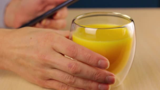 Glas met sinaasappelsap op de voorgrond. De vrouw vingers raken het scherm van de smartphone op de achtergrond en pakt periodiek een kopje drank met zijn hand. - Video