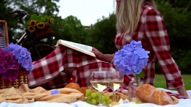Dívka v červených kostkovaných šatech a klobouku sedí na bílém pleteném piknikovém prostěradle a čte knihu a pije víno. Letní piknik za slunečného dne s chlebem, ovocem, kyticemi a hortenzií. Selektivní zaměření - Záběry, video