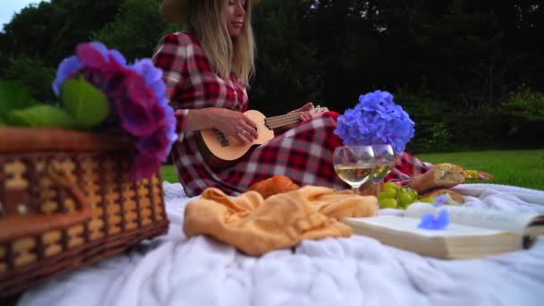 Chica en vestido rojo a cuadros y sombrero sentado en blanco punto manta de picnic juega ukelele y beber vino. Picnic de verano en el día soleado con pan, fruta, flores de hortensias ramo. Enfoque selectivo - Metraje, vídeo