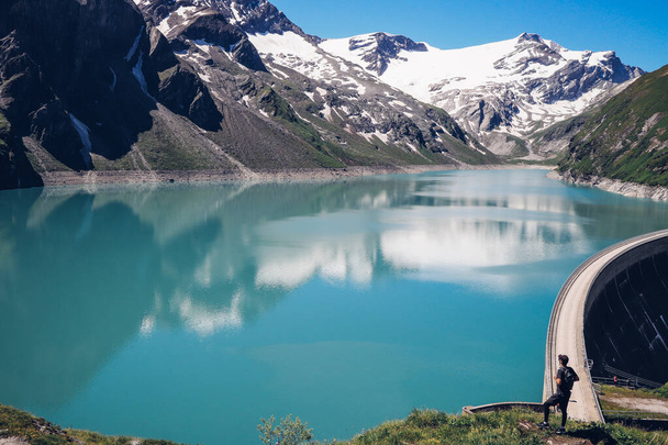 Vue sur le barrage du glacier Stausee Mooserboden, qui s'élève sous le glacier Kaprun et est l'un des plus grands producteurs d'électricité d'Autriche. Le garçon intrépide se tient au bord du rocher et admire. - Photo, image