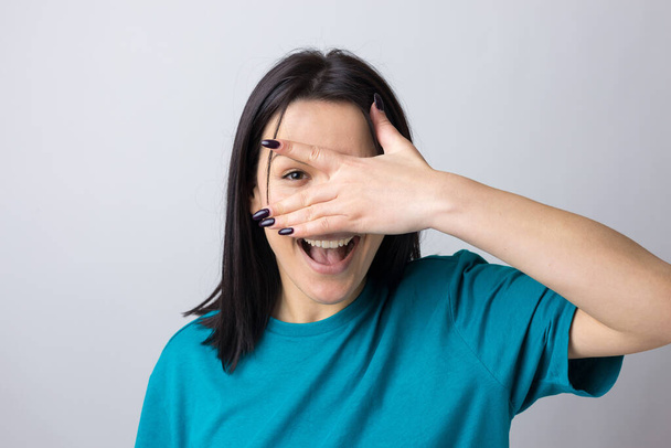 Picture of brunette teenage girl covering face with hands and peeping through her fingers. - Photo, Image