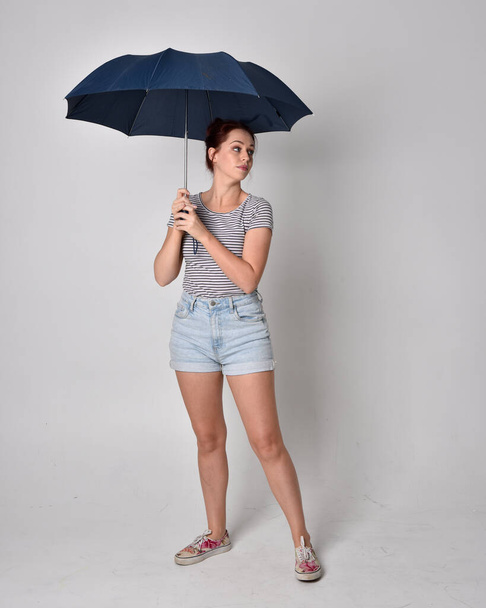 retrato de longitud completa de la niña con camisa y pantalones cortos de mezclilla, sosteniendo un paraguas. aislado sobre fondo gris estudio. - Foto, imagen