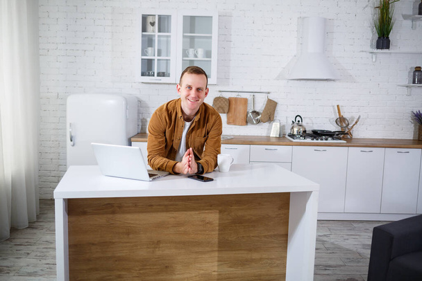 Schöner junger Geschäftsmann, der mit dem Computer arbeitet und in der Küche am Tisch sitzt. Ein angenehmer, glücklicher Mensch kommuniziert in einem sozialen Netzwerk, sucht Informationen im Internet. - Foto, Bild