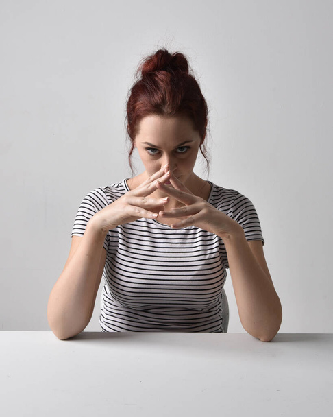 3/4 close up portrait of red haired girl wearing striped shirt. Leaning on table pose with studio background. - Photo, Image