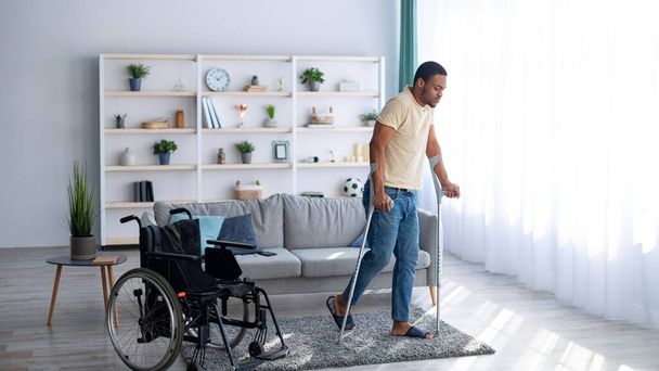 Conceito de reabilitação. Jovem negro caminhando com muletas depois de usar cadeira de rodas em casa, panorama - Foto, Imagem
