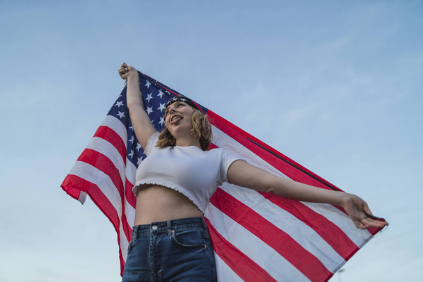 A young Caucasian female holding the flag of the USA against the blue sky - Foto, immagini