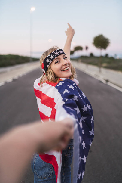 A smiling young Caucasian female holding the hand of her friend with the USA flag on her shoulders - Foto, immagini