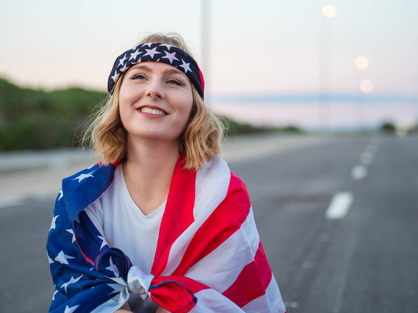 A patriotic Caucasian woman with a nose ring and a trendy bandana sitting in the middle of the road with the US flag draped around her - Foto, Bild