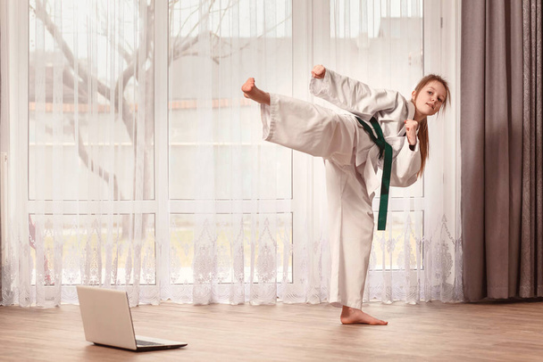 Kimonolu kız dövüş pozisyonunda duruyor. Çocuk evde dizüstü bilgisayarın önünde karate eğitimi veriyor. Tekvando dövüşçüsü tekmeler.. - Fotoğraf, Görsel