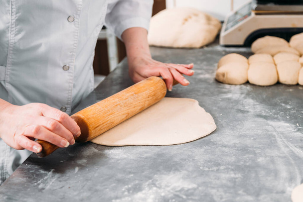 Female hands roll out a sheet of dough with a rolling pin on the table next to a piece of dough and a baking dish.  - Foto, Imagem