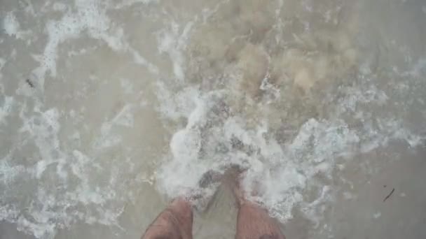 Çıplak ayaklı bir adamın çıplak ayakla sahilin üzerinde köpüklü dalgalarla duran görüntüsü. - Video, Çekim