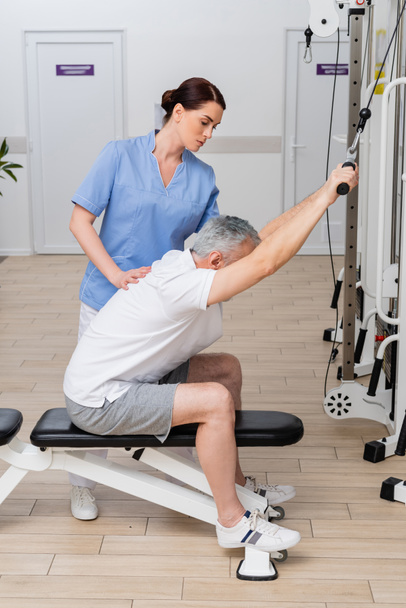 rehabilitólogo ayudando a hombre maduro a hacer ejercicio en el gimnasio del hospital - Foto, Imagen
