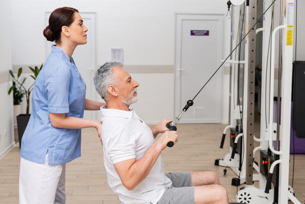 vista lateral de reabilitologist que apoia o homem maduro que exercita-se na máquina do cabo da tração - Foto, Imagem