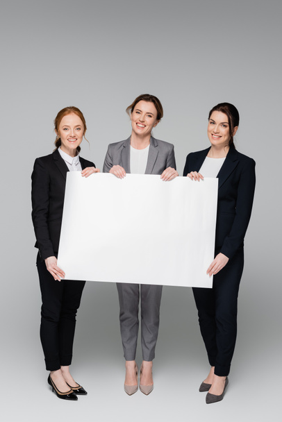 Cheerful businesswomen with white placard on grey background - Photo, Image