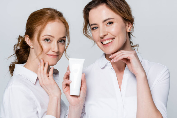 Smiling women looking at camera while holding tube with cream isolated on grey  - Photo, Image