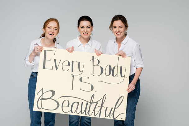Cheerful women holding placard with every body is beautiful lettering isolated on grey  - Photo, Image