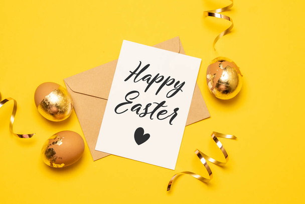 Bonne composition de Pâques avec du papier blanc et du texte de bienvenue, des œufs d'or et de la serpentine sur fond jaune vif - Photo, image