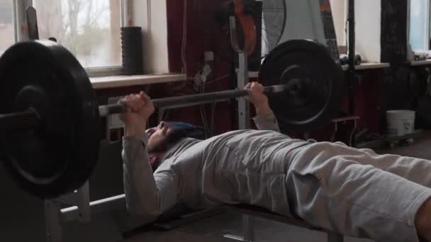 Młody mężczyzna napinający mięśnie, trenujący sztangę we wnętrzu siłowni - Materiał filmowy, wideo