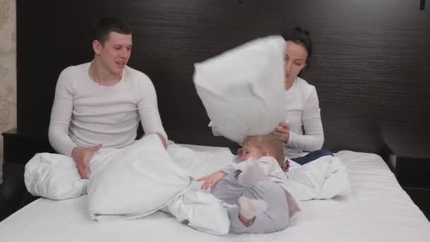 Happy family, child, daughter, mom, dad, play together with pillows, in room on the bed. Young parents are playing with a small child. Kid, mom, father, pillow fight game. Family laughs while playing - Footage, Video