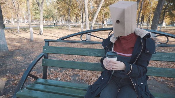 La chica con el paquete en la cabeza sentada en un banco con una taza de café. Chica en una chaqueta - Foto, Imagen