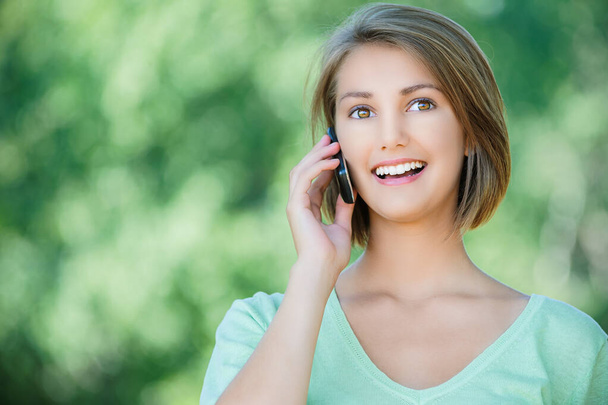 Młoda słodka śmiejąca się kobieta w zielonej bluzce rozmawiająca przez telefon komórkowy w letnim parku. - Zdjęcie, obraz