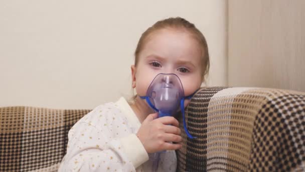 Behandeling van een kind thuis voor griep en verkoudheid van de longen. Het kind wordt ingeademd met een vernevelaar terwijl het op de bank in de kinderkamer zit. Een klein meisje ademt in een masker met medicijnen - Video