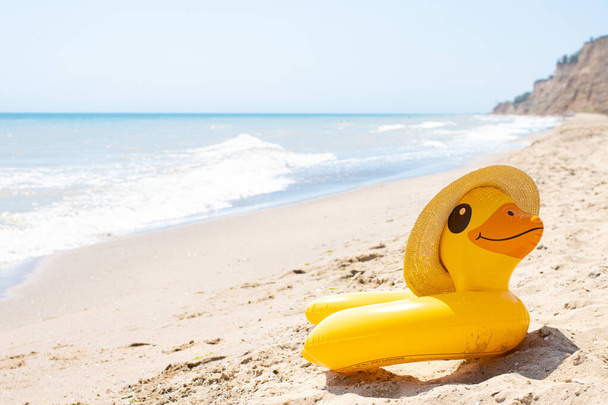 Anello gonfiabile giallo dell'anatra nel cappello di paglia di estate che posa sulla spiaggia vuota sabbiosa vicino all'oceano ondulato blu nella giornata soleggiata. Tubo di protezione per bambini. Concetto di villeggiatura estiva. Copia spazio - Foto, immagini