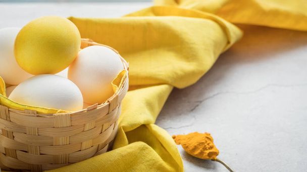 Gekleurde paaseieren en witte eieren in een mandje op een gele doek met kopieerruimte. Geel paasei gekleurd door kurkuma poeder .Natuurlijke kleurstof voor eieren. Paaseieren beschilderd met natuurlijke kleurstoffen . - Foto, afbeelding