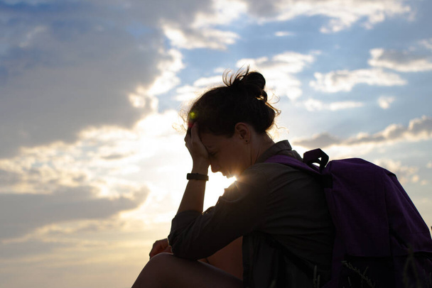 ハイキングの冒険の間に休んで座ってバックパックを持つ疲れた女性ハイカーのシルエット。日没の光と曇りの空の背景に旅行者の側のビュー。目標達成の困難を克服する. - 写真・画像